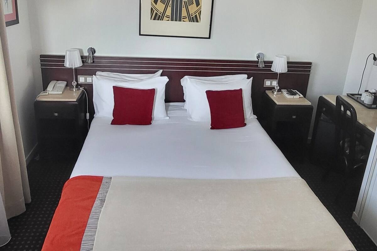 Hotelkamer met een groot bed, twee nachtkastjes, een bureau met stoel, een airconditioner en een lijst aan de muur.