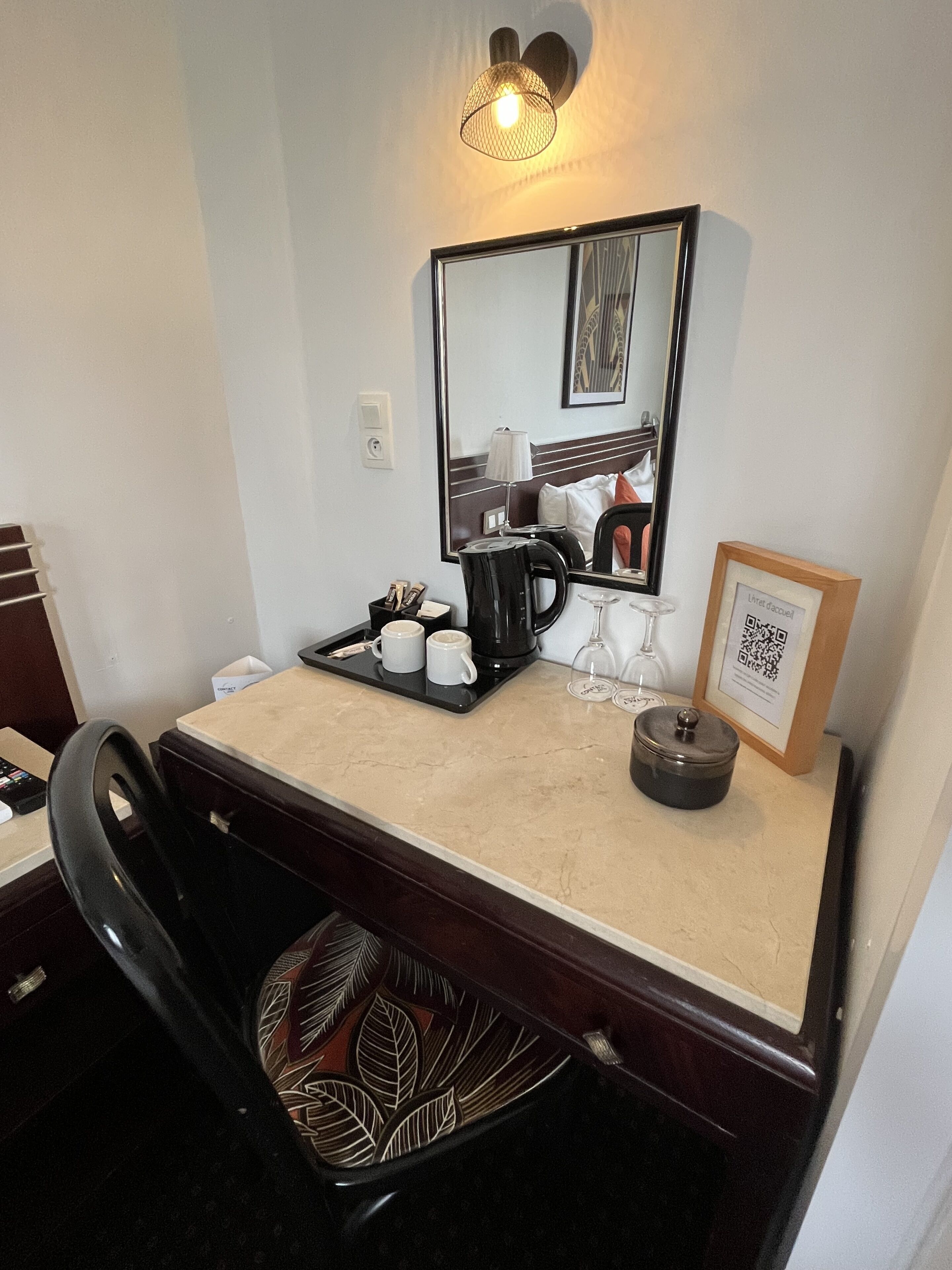 Una habitación de hotel con escritorio, una silla, cafetera, bandeja con tazas, copa de vino, cuadro enmarco y espejo.
