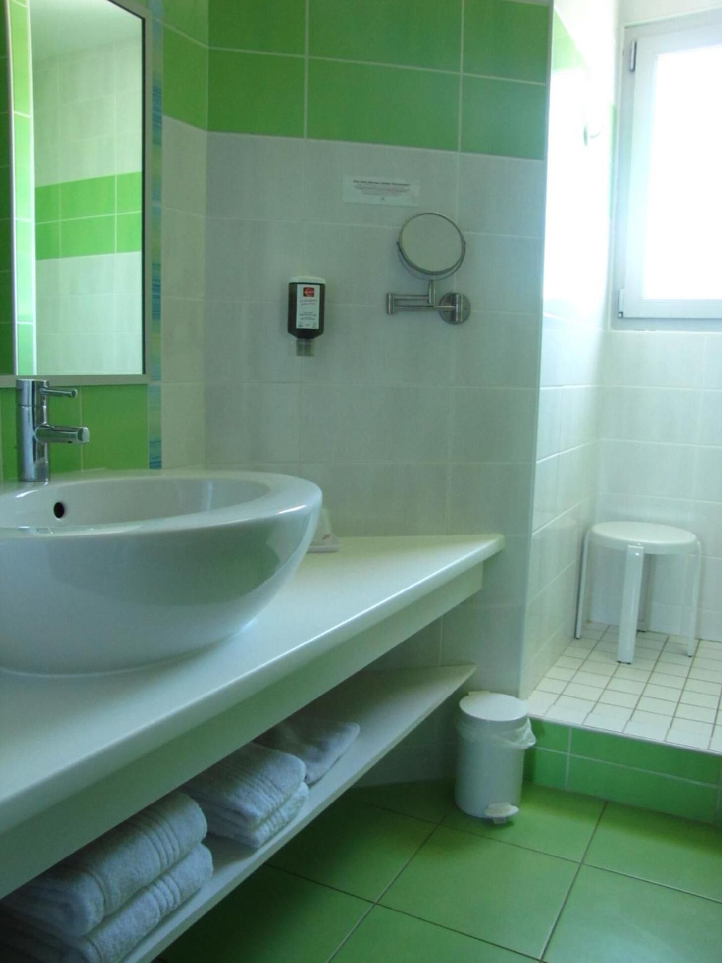 A bathroom with a white sink, green tiles, a mirror, a soap dispenser, a small trash bin, and a window.