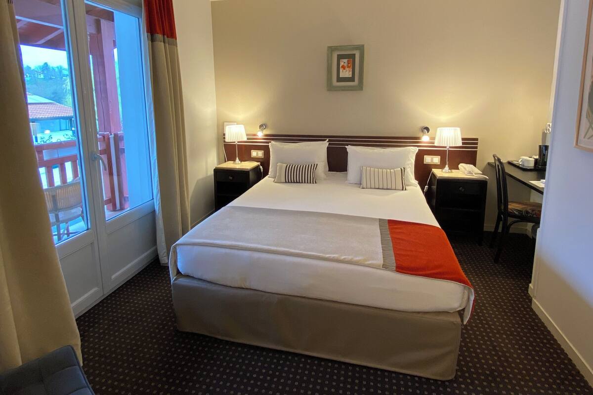 Een hotelkamer met een groot bed, twee nachtkastjes met lampen, een bureau, een stoel, een raam met gordijnen en een ingelijst kunstwerk aan de muur.