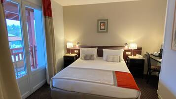 A hotel room with a large bed, two bedside tables with lamps, a desk, a chair, a window with curtains, and a framed picture on the wall.