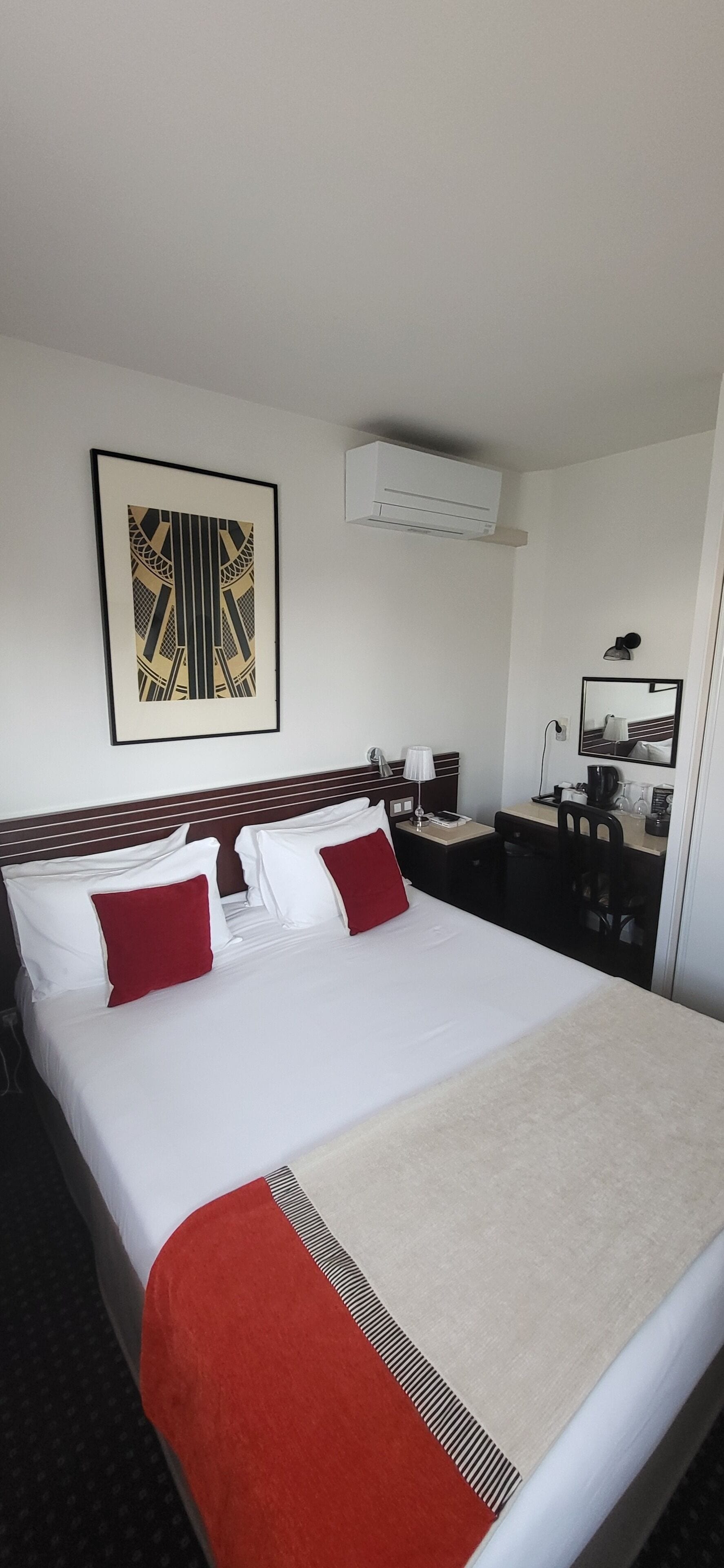 Quarto de hotel com cama, mesa de cabeceira, escrivaninha, cadeira, espelho, ar condicionado e um quadro na parede.