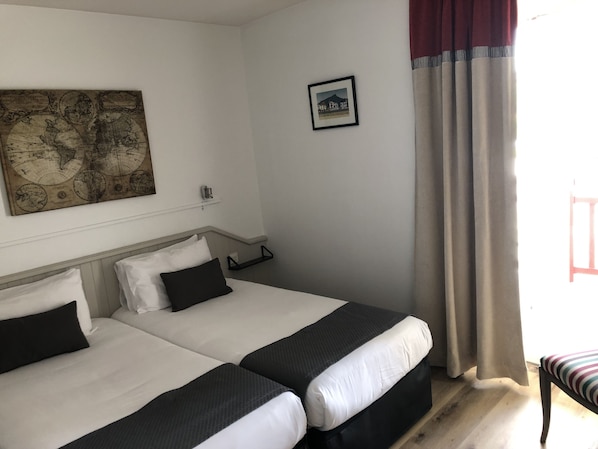 A hotel room with two beds, a world map on the wall, a framed picture, and a window with curtains.