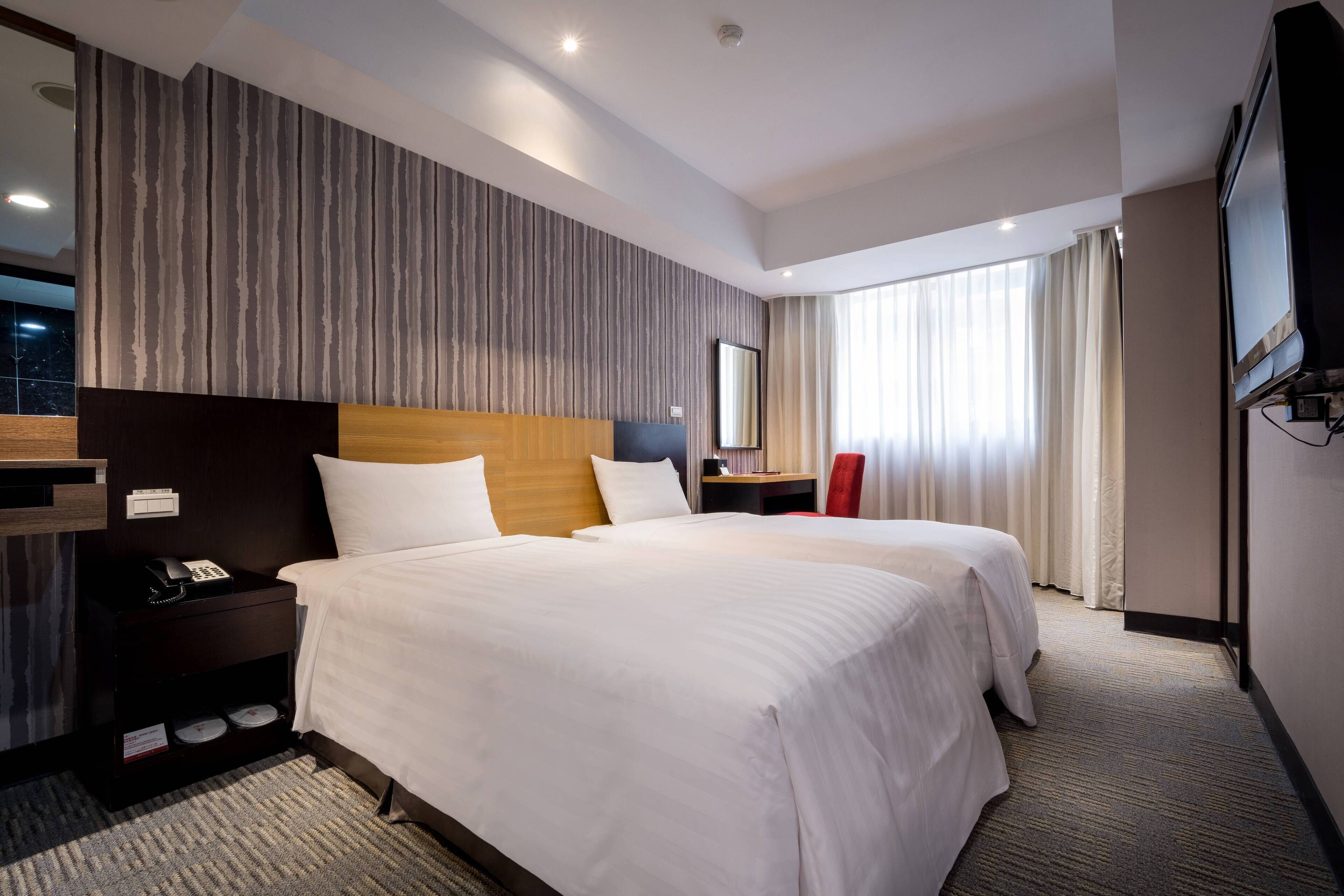 A hotel room with two beds, a wooden headboard, a nightstand with a phone, a mirror, and a TV.