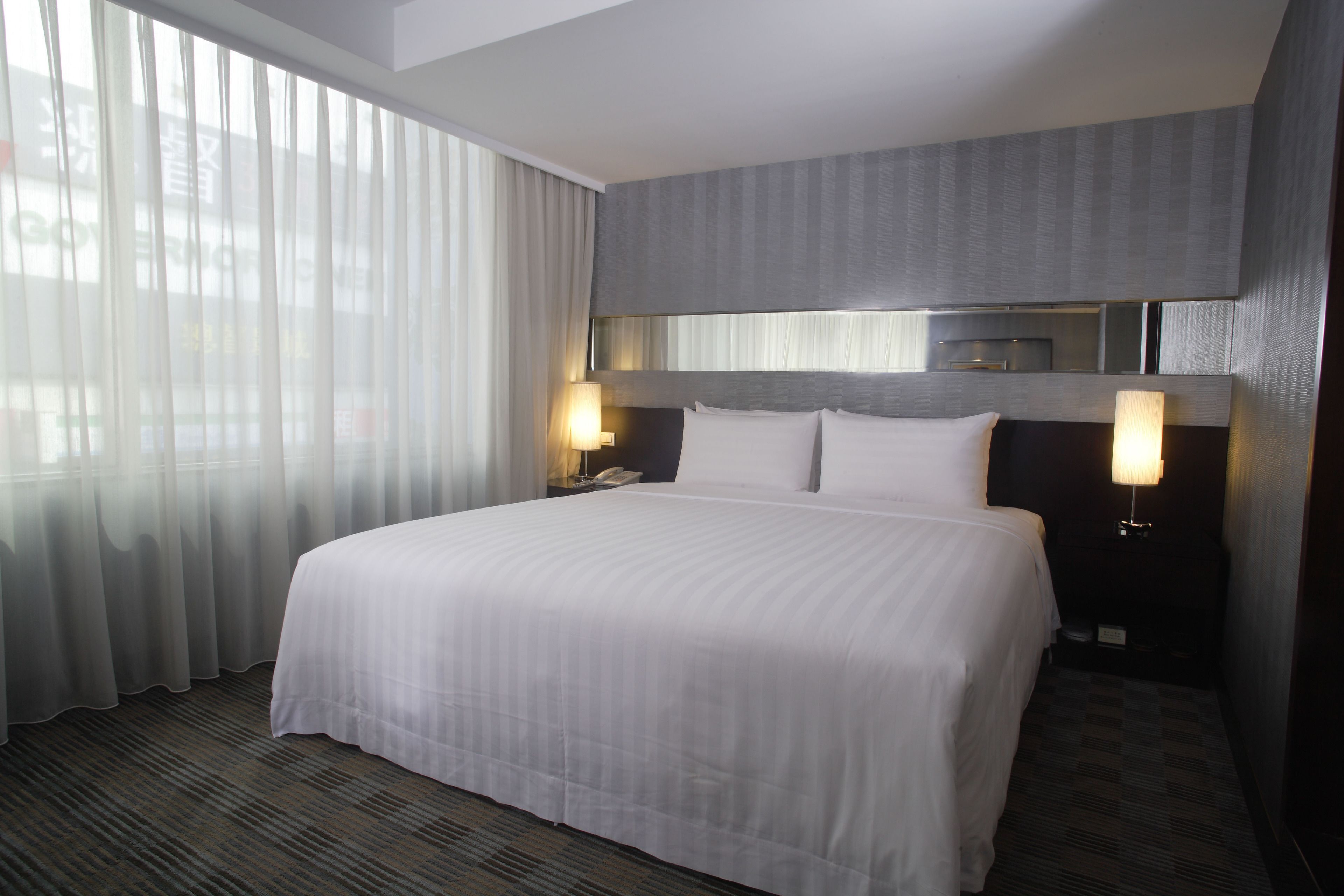 A hotel room with a large bed, two bedside tables with lamps, a headboard, and a window with sheer curtains.