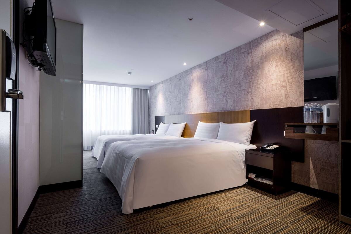 Habitación de hotel con una cama grande, una mesita de noche con teléfono, un televisor de pantalla plana montado en la pared y una ventana con cortinas.