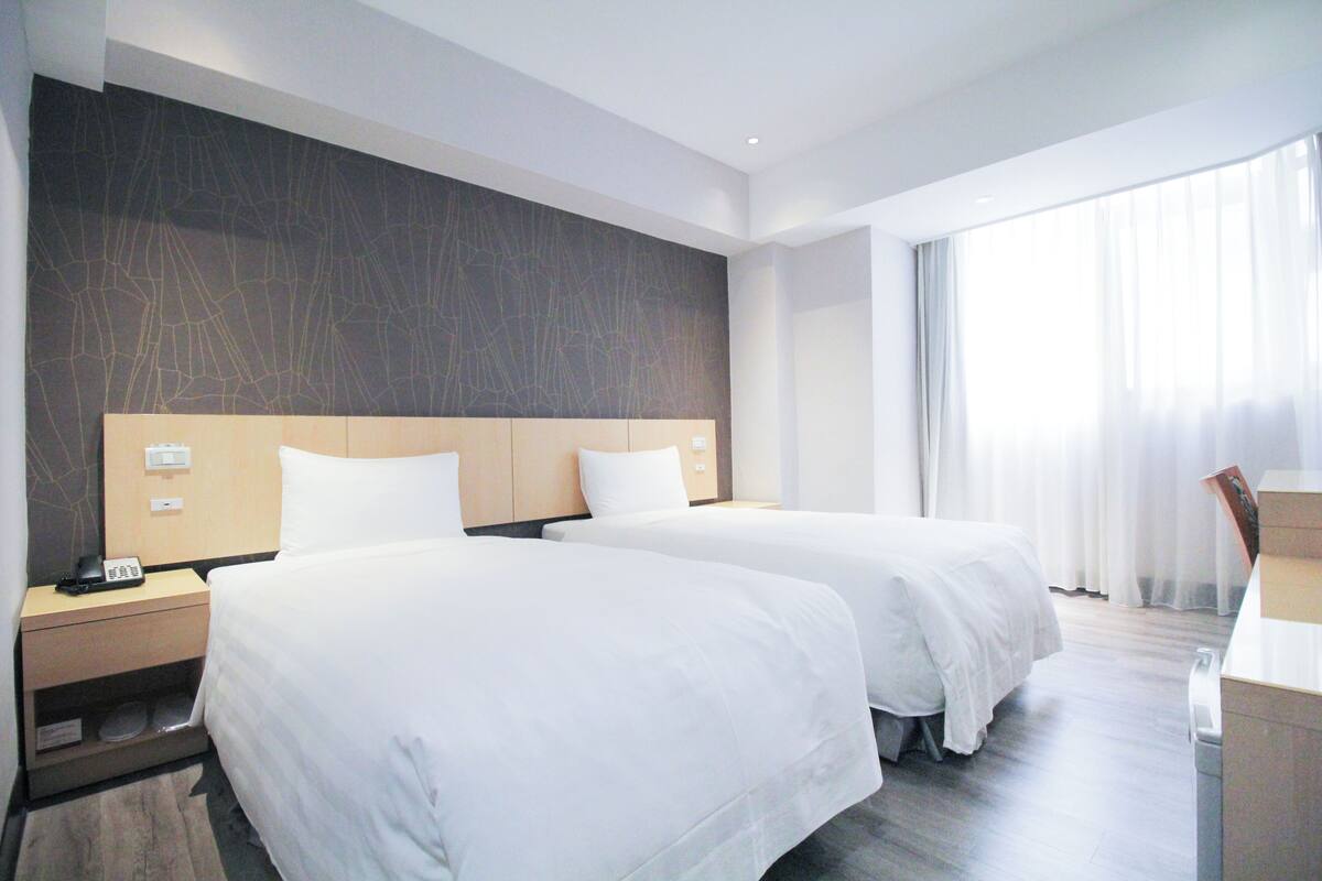 Habitación de hotel con dos camas, cabecero de madera, mesita de noche con teléfono y un ventanal con cortinas.