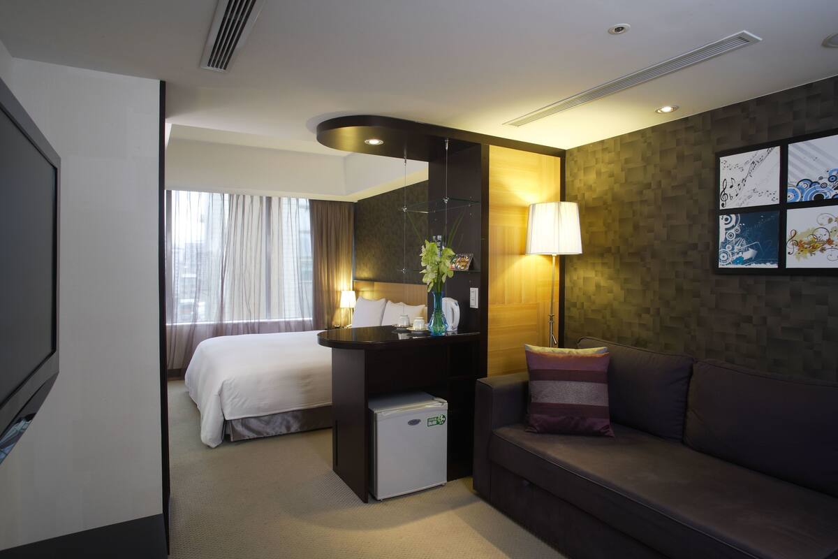 Una habitación de hotel con cama, un pequeño escritorio con lámpara, un sofá y una pared decorada con obras de arte.