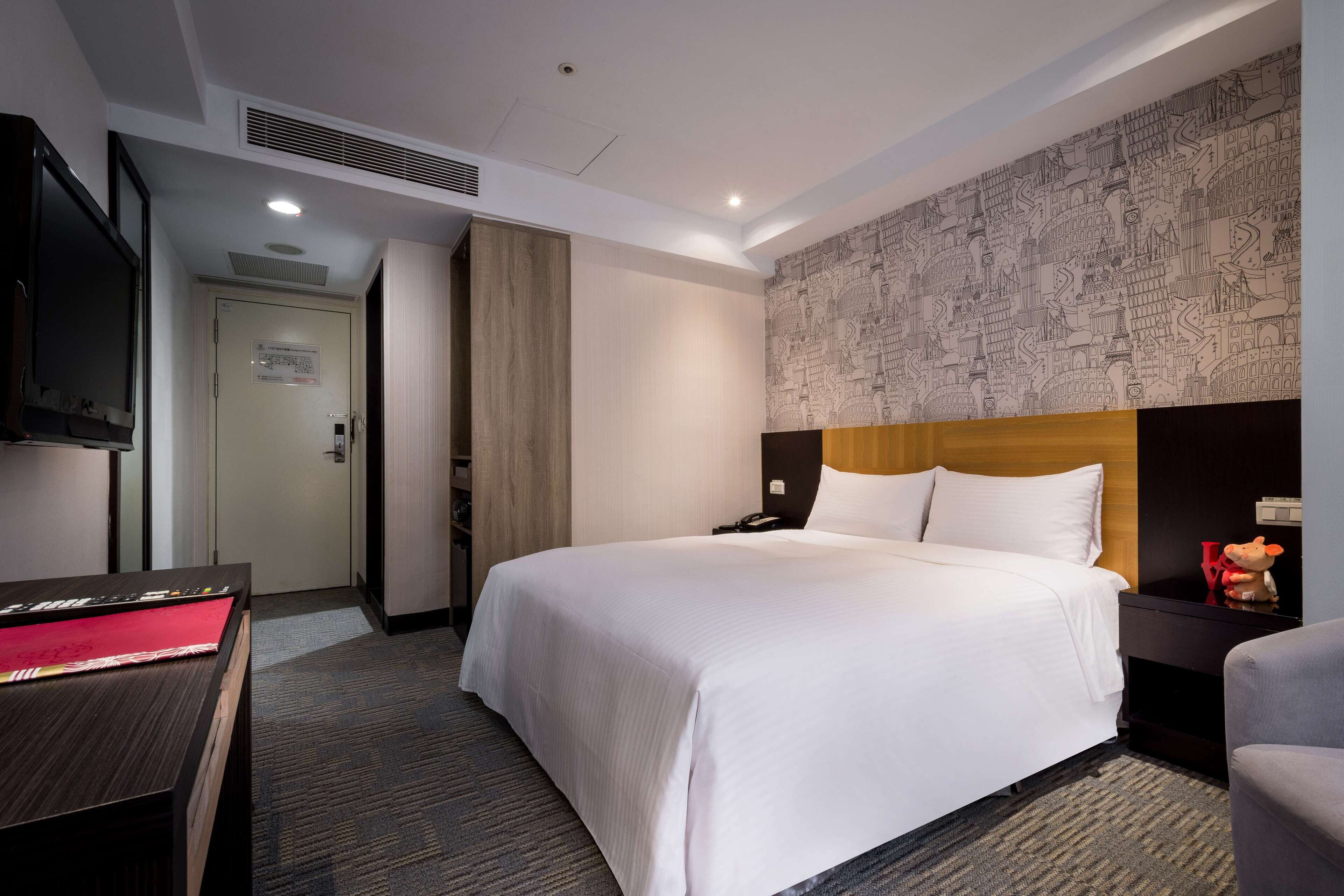 Habitación de hotel con una cama grande, un escritorio con televisor de pantalla plana, un cabecero de madera y una pared con un mural de un paisaje urbano.