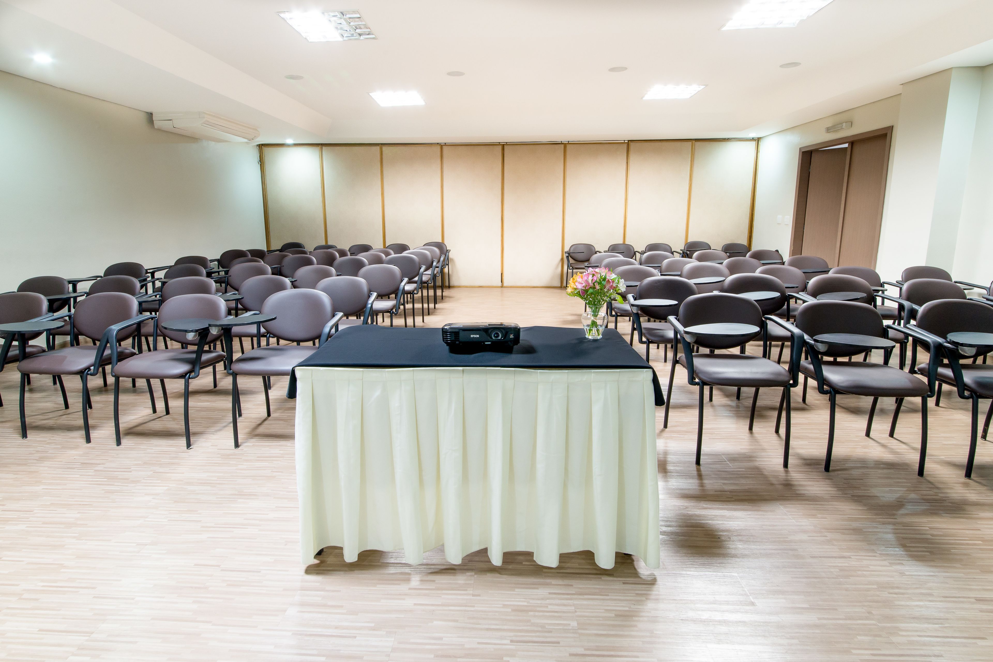 Un salón de conferencias con filas de sillas mirando hacia un escenario, una mesa con un micrófono y un jarrón con flores.