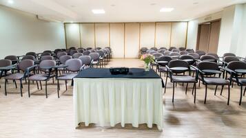Un salón de conferencias con filas de sillas mirando hacia un escenario, una mesa con un micrófono y un jarrón con flores.
