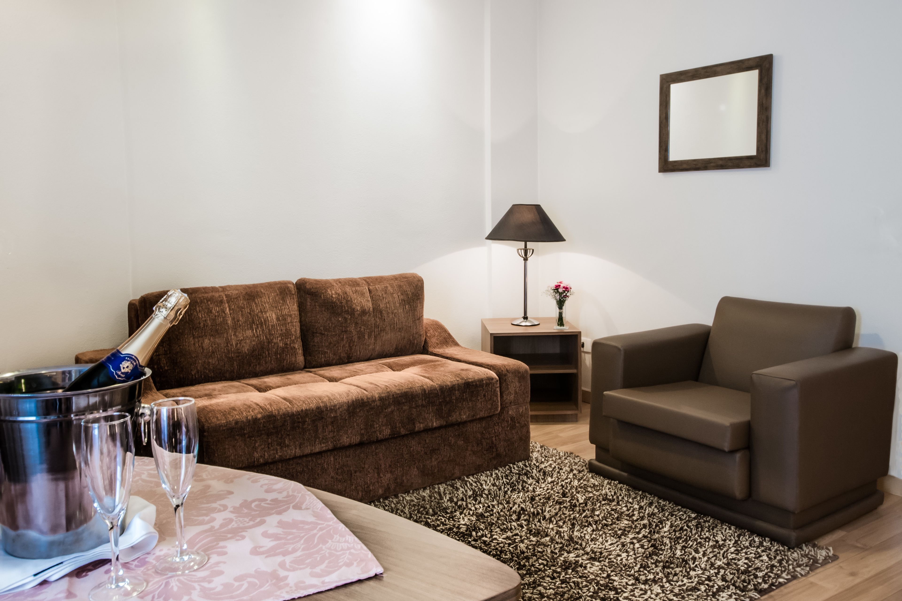 A living room with a brown sofa, a brown armchair, a small table with a bottle of champagne and glasses, a lamp, and a framed picture on the wall.