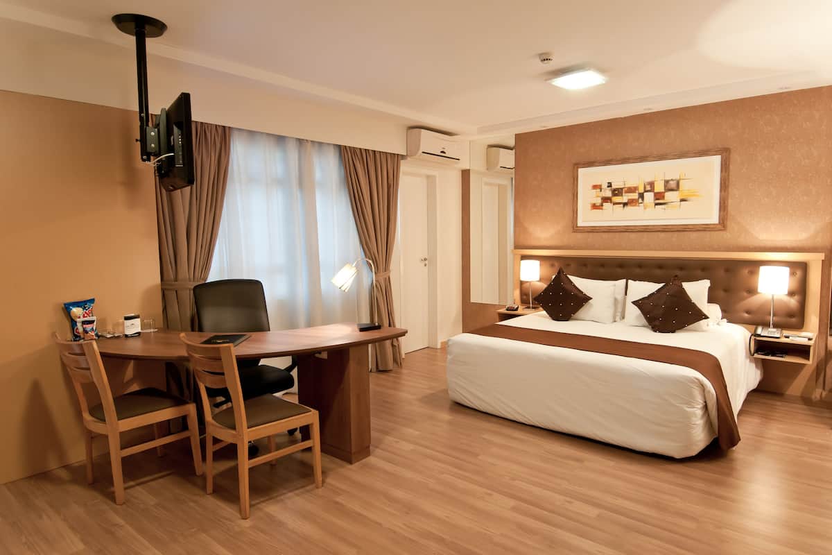 A hotel room with a bed, a desk, and a chair. There is a television mounted on the wall, an air conditioning unit, and a framed picture on the wall.