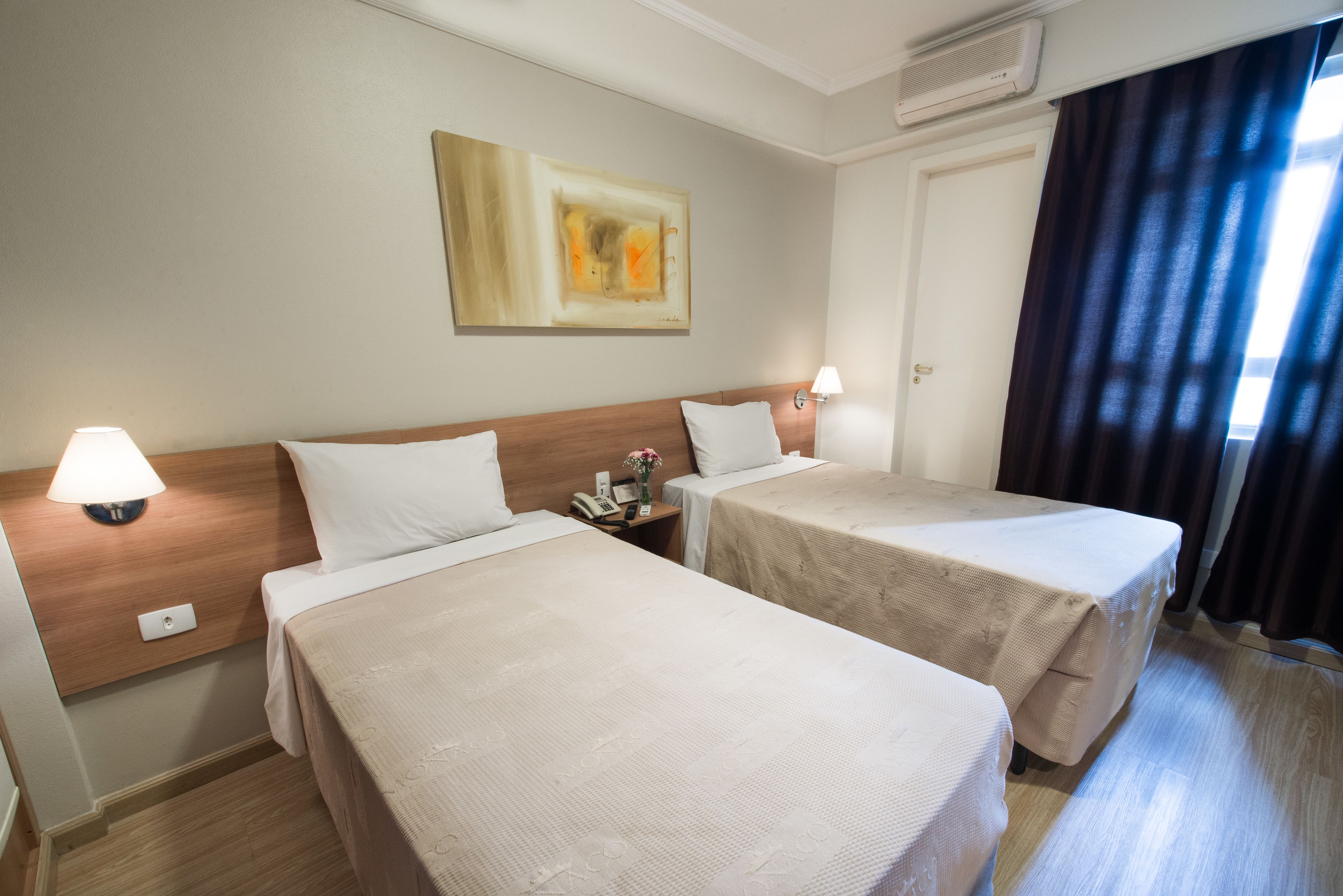 A hotel room with two single beds, a wall-mounted artwork, a bedside table with a lamp, and a window with curtains.