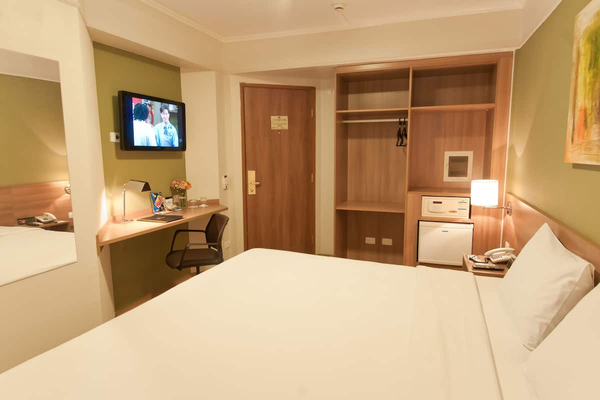 A hotel room with a bed, a desk with a chair, a television, a wardrobe, and a small refrigerator.
