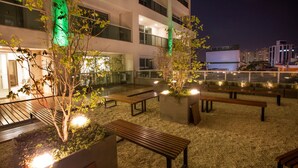 Un área de estar al aire libre con bancos y macetas, rodeada por un edificio con columnas iluminadas.