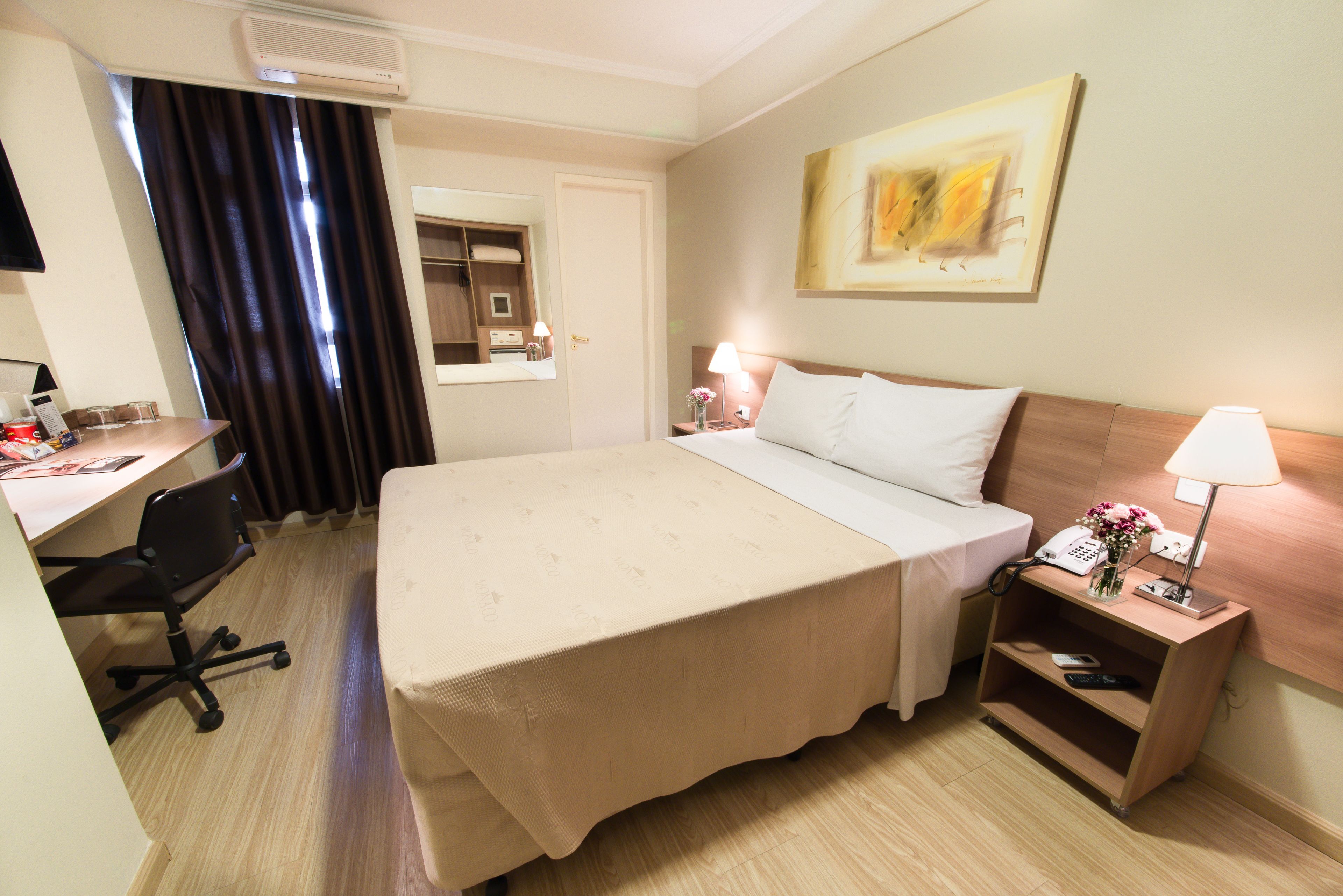 A hotel room with a bed, desk, chair, bedside table with lamp, and a painting on the wall.