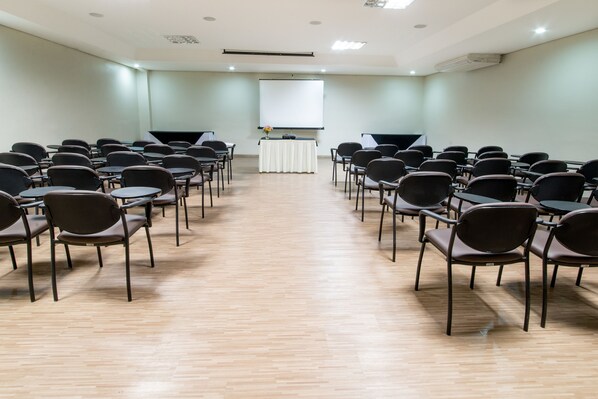 A conference room with rows of chairs facing a white screen, a table with a vase of flowers, and a projector.