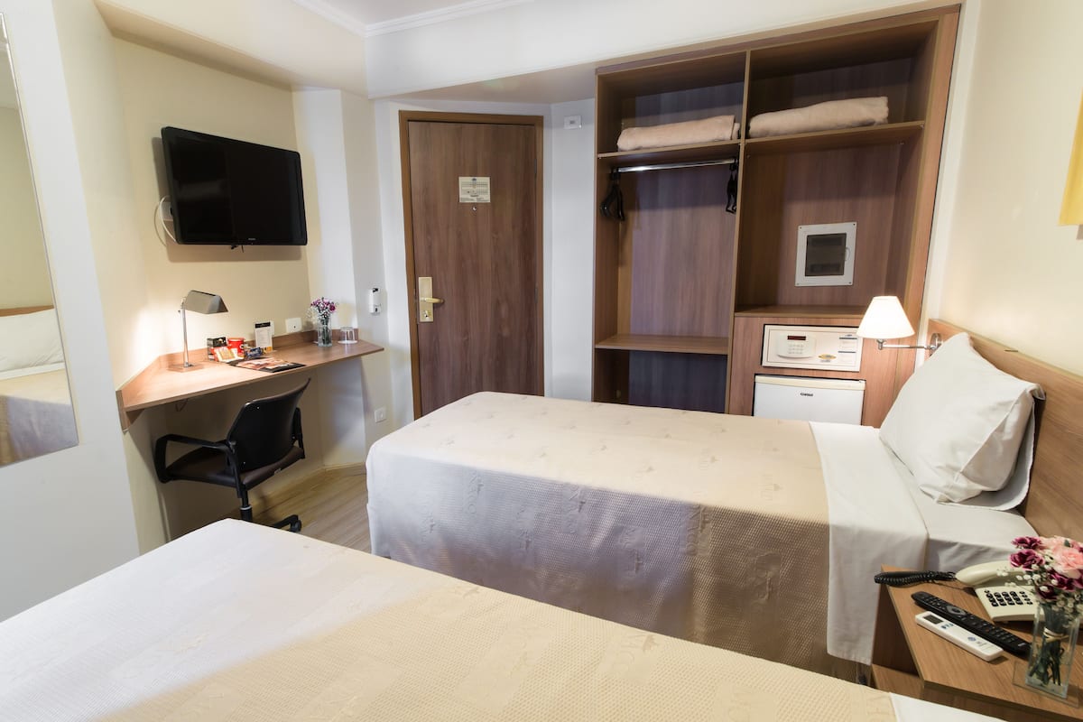 A hotel room with two beds, a desk, a TV, a wardrobe, and a bedside table with a lamp and phone.