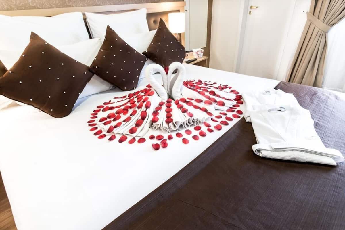 A neatly made bed with a swan-shaped arrangement of red rose petals, white pillows, and a folded towel.