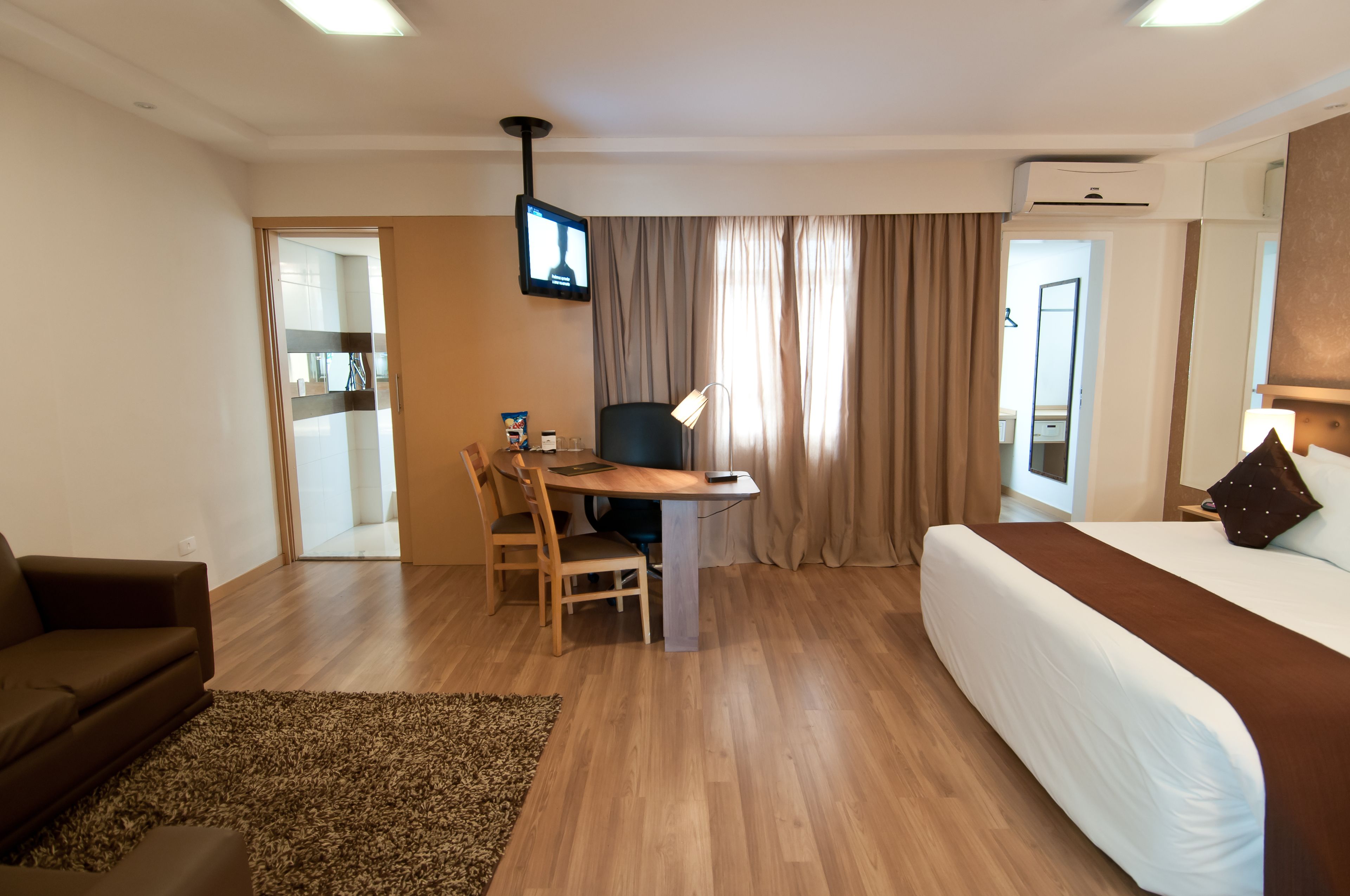 A hotel room with a bed, a desk with a chair, a TV mounted on the wall, and a window with curtains.