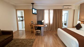 A hotel room with a bed, a desk with a chair, a TV mounted on the wall, and a window with curtains.