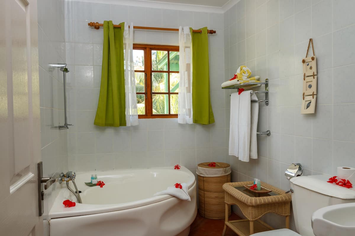 A bathroom with a bathtub, a toilet, a sink, a window with curtains, and a small wicker basket with toiletries.