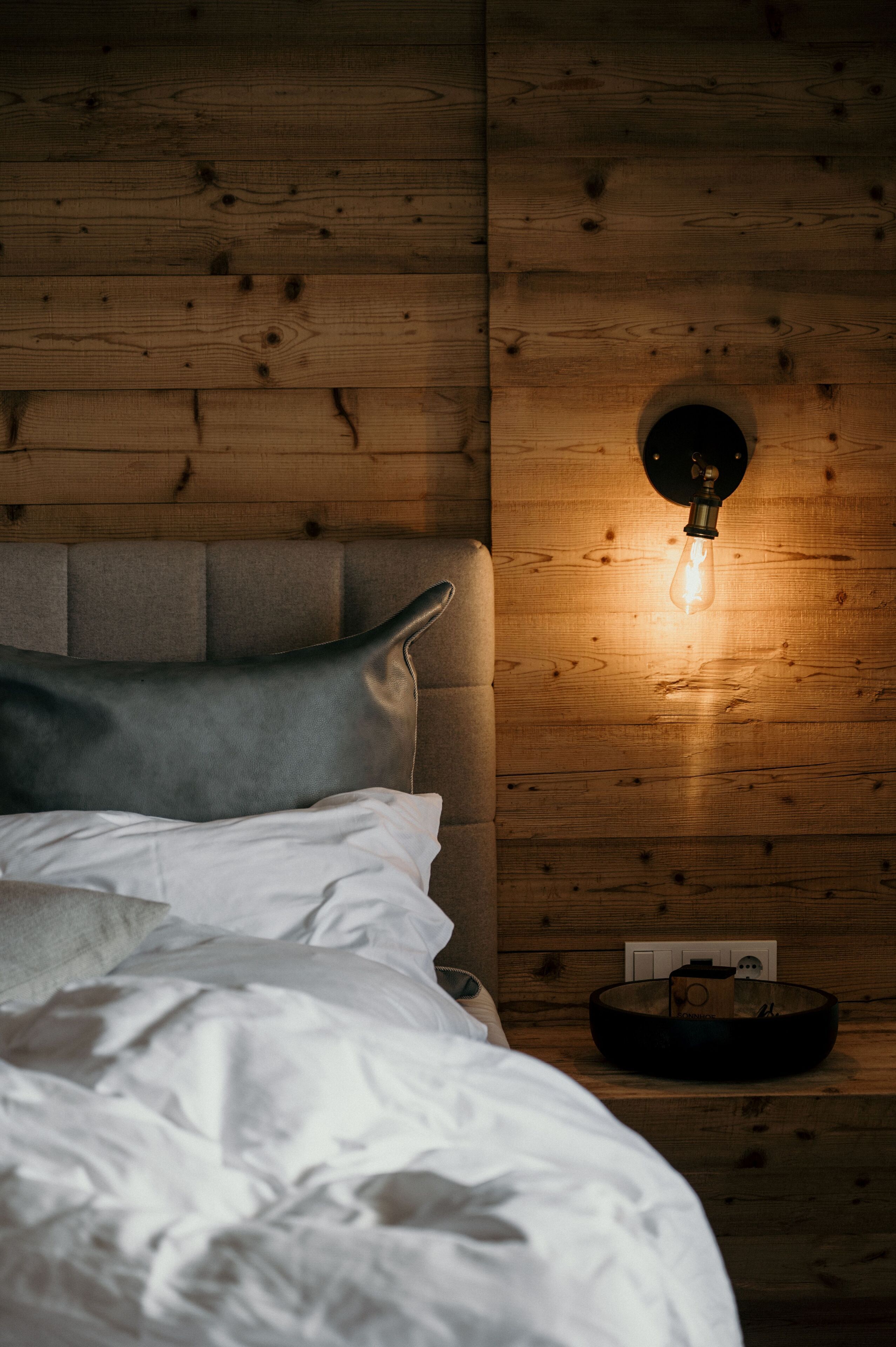 A bedroom with a wooden headboard, a bedside lamp, and a small table with a bowl and a box.