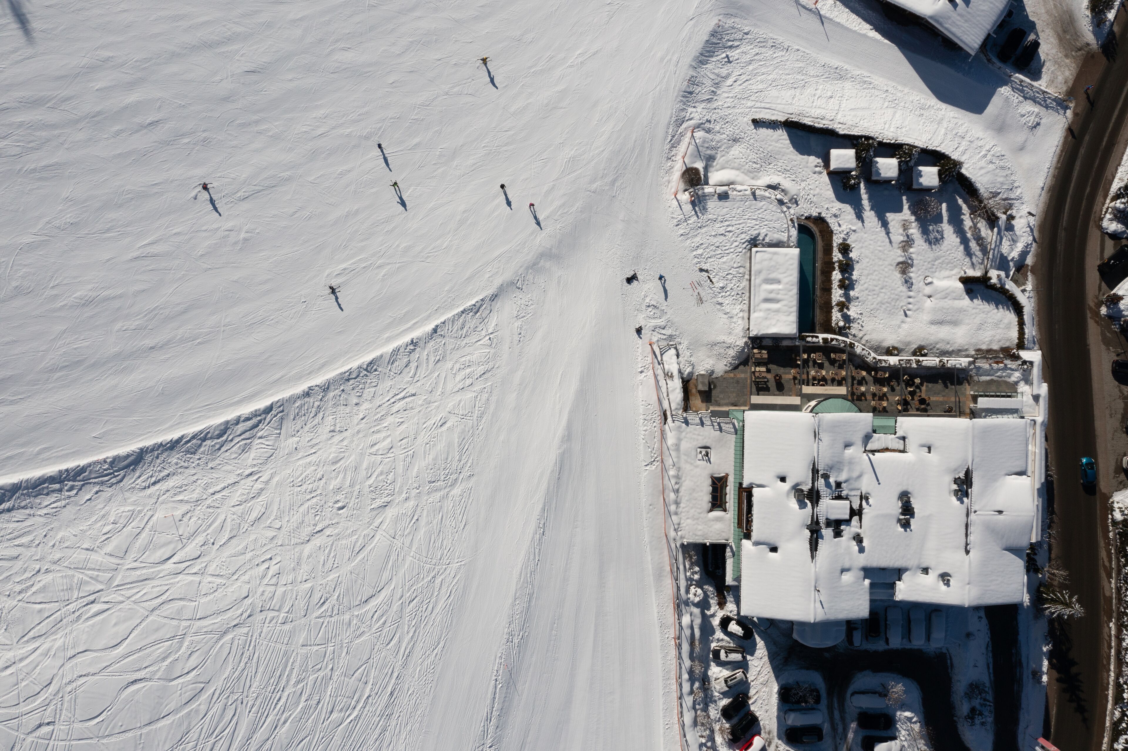 An aerial view of a snow-covered area with ski tracks, a parking lot, and a building with multiple windows and a green roof.