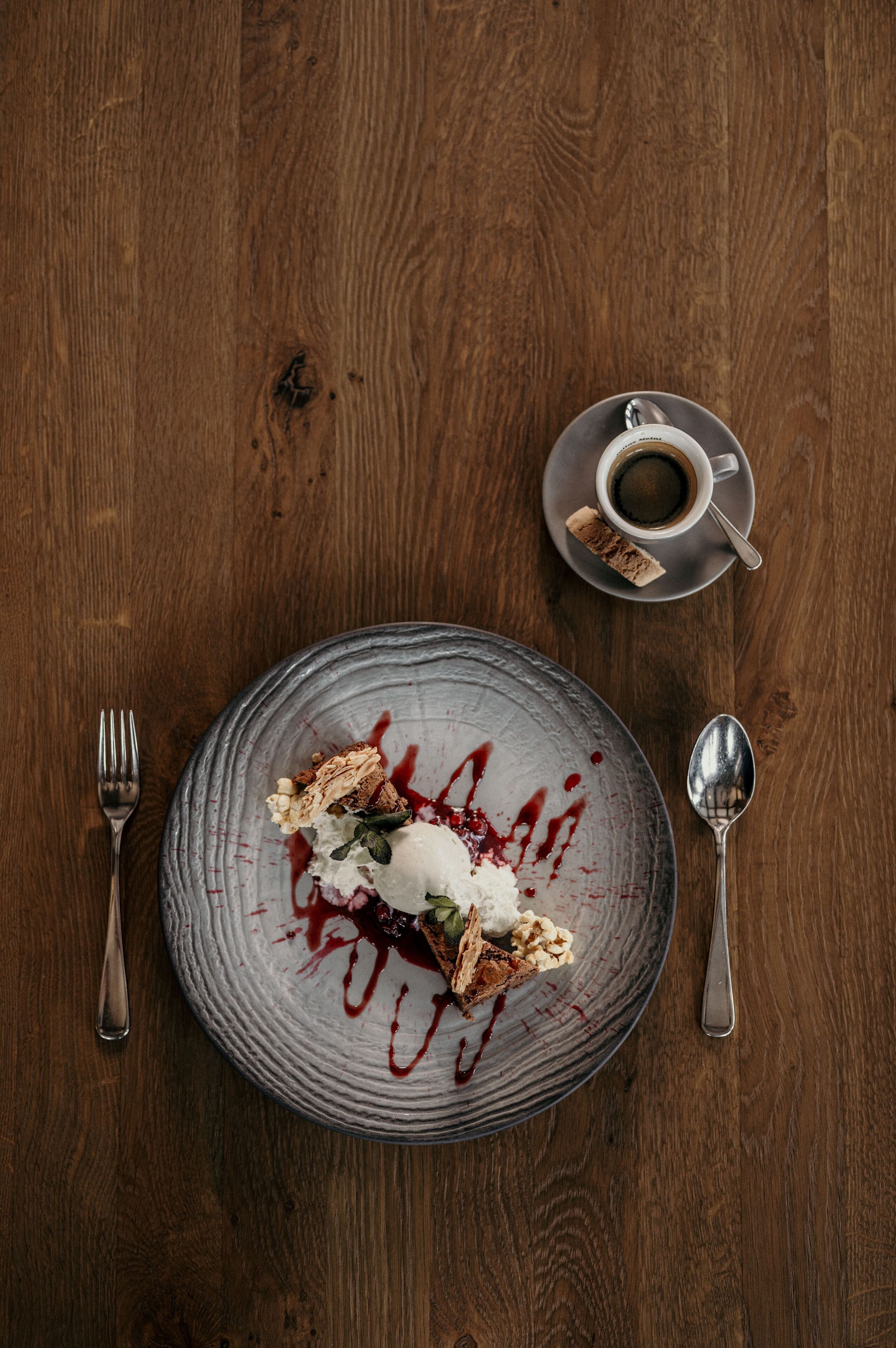Een nagerechtenbord met een bolletje ijs en bessen op een gestructureerd bord, samen met een kop koffie en een klein taartje op een schoteltje.