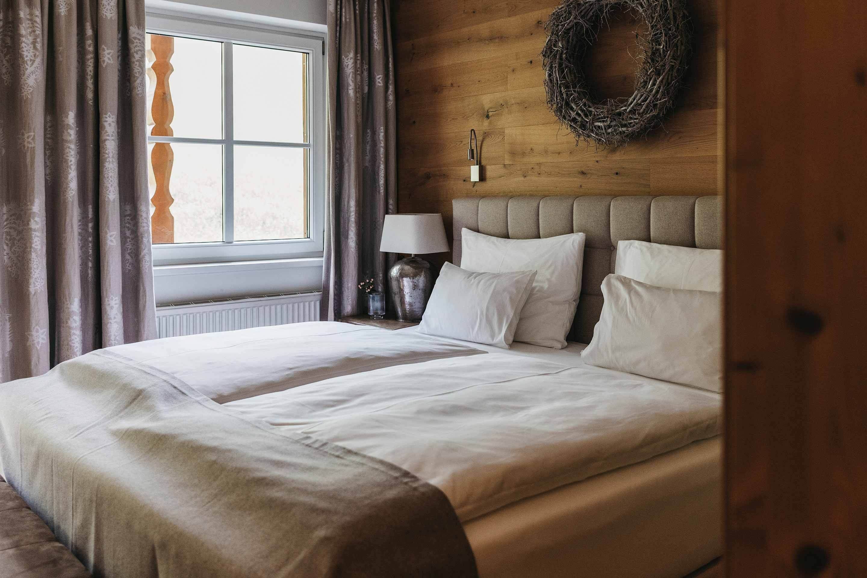 A neatly made bed with white linens, a wooden headboard, a bedside lamp, and a window with curtains.