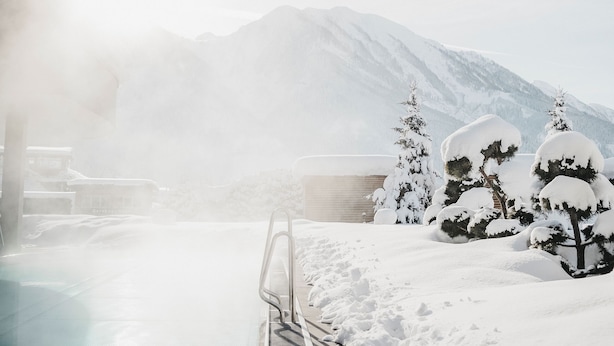 A snow-covered outdoor pool area with a metal railing, surrounded by snow-laden trees and mountains in the background.