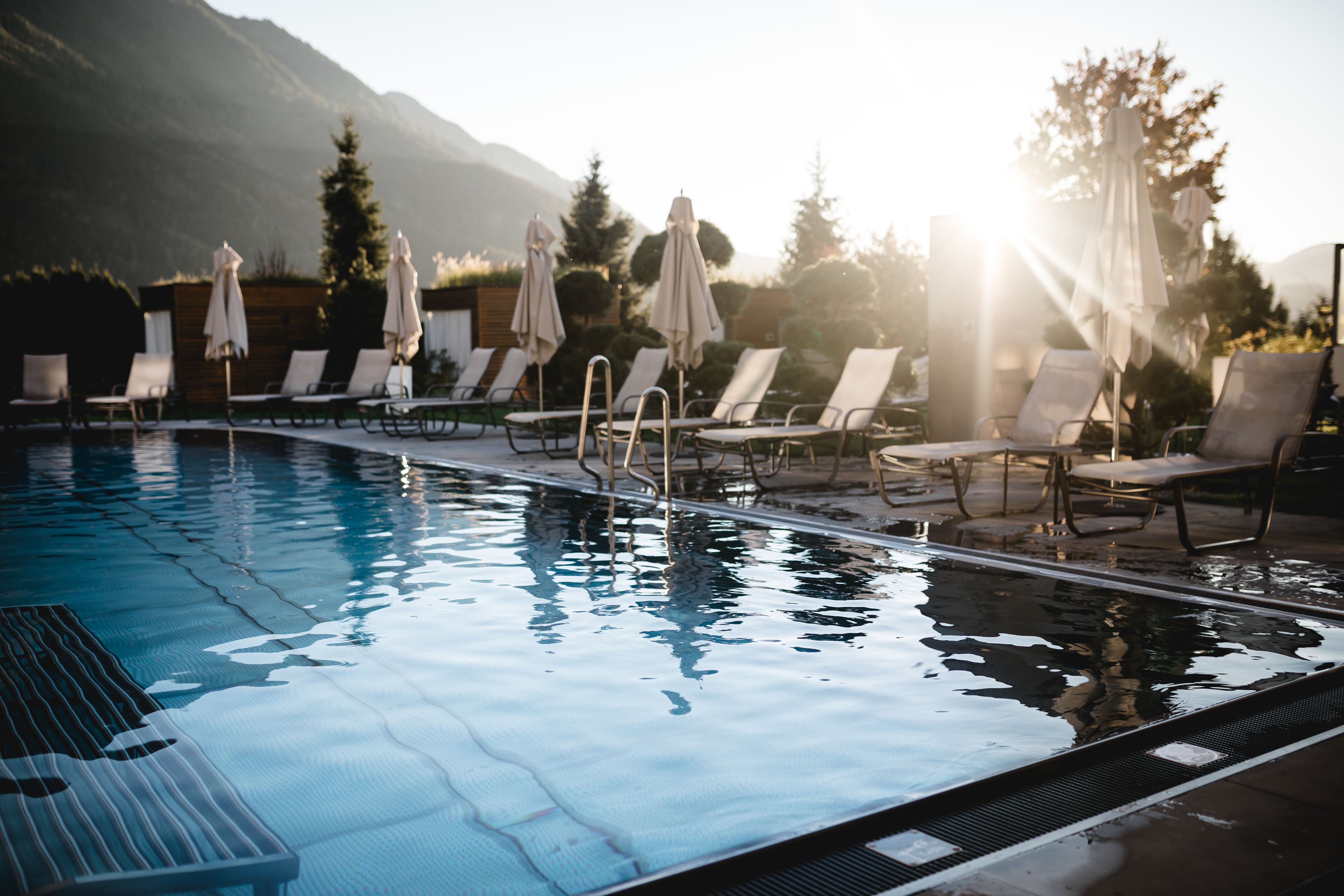Una piscina con lettini e ombrelloni, circondata da alberi e montagne.
