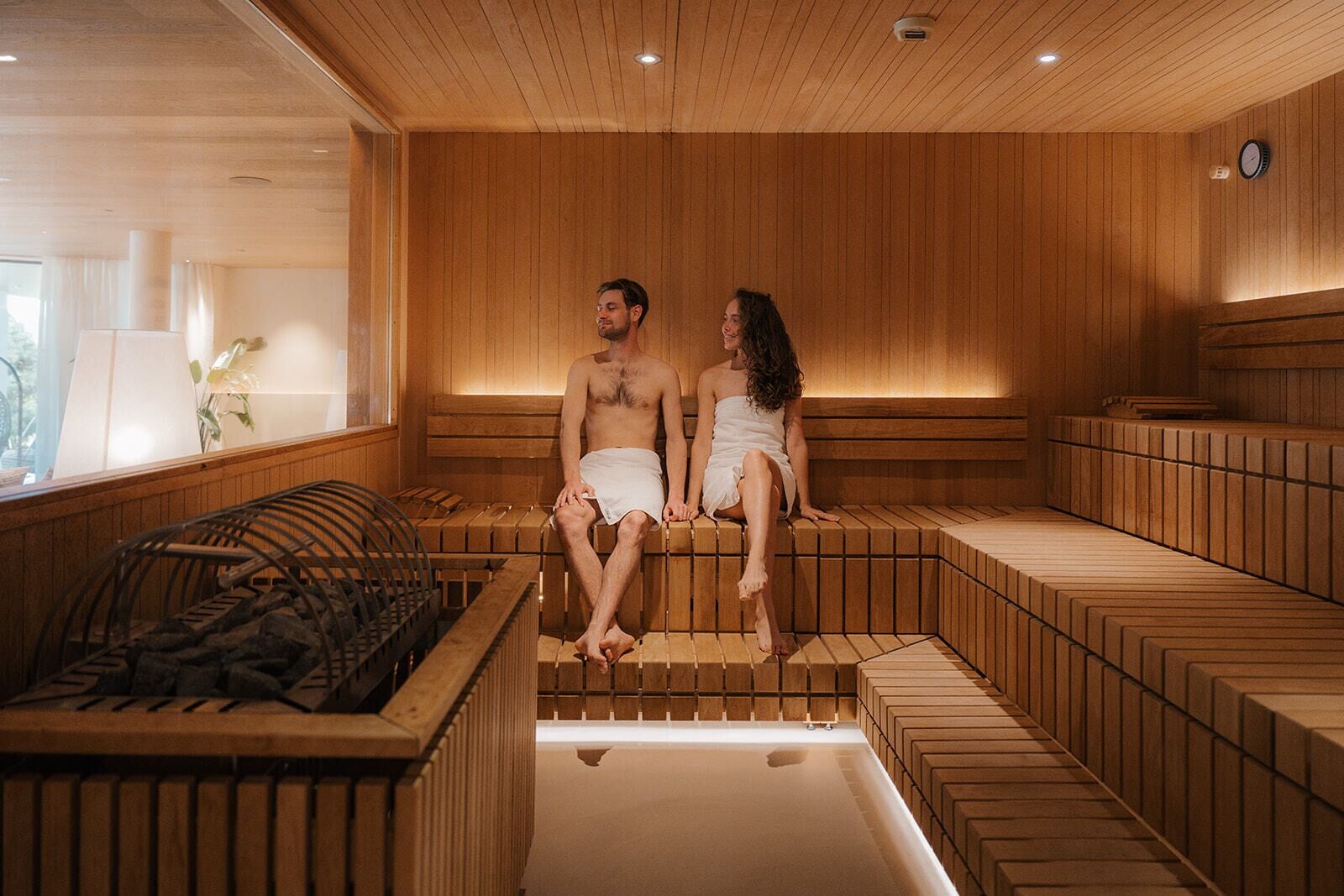 Una sauna con bancos de madera y suelo calefaccionado, una ventana con vistas y un reloj en la pared.