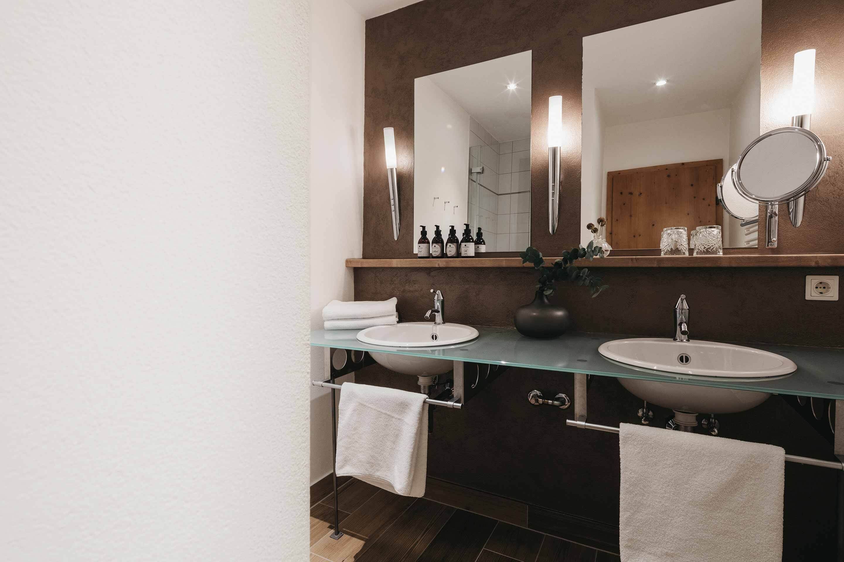 Une salle de bain moderne avec deux lavabos, un grand miroir et une étagère en verre où sont rangés les articles de toilette.