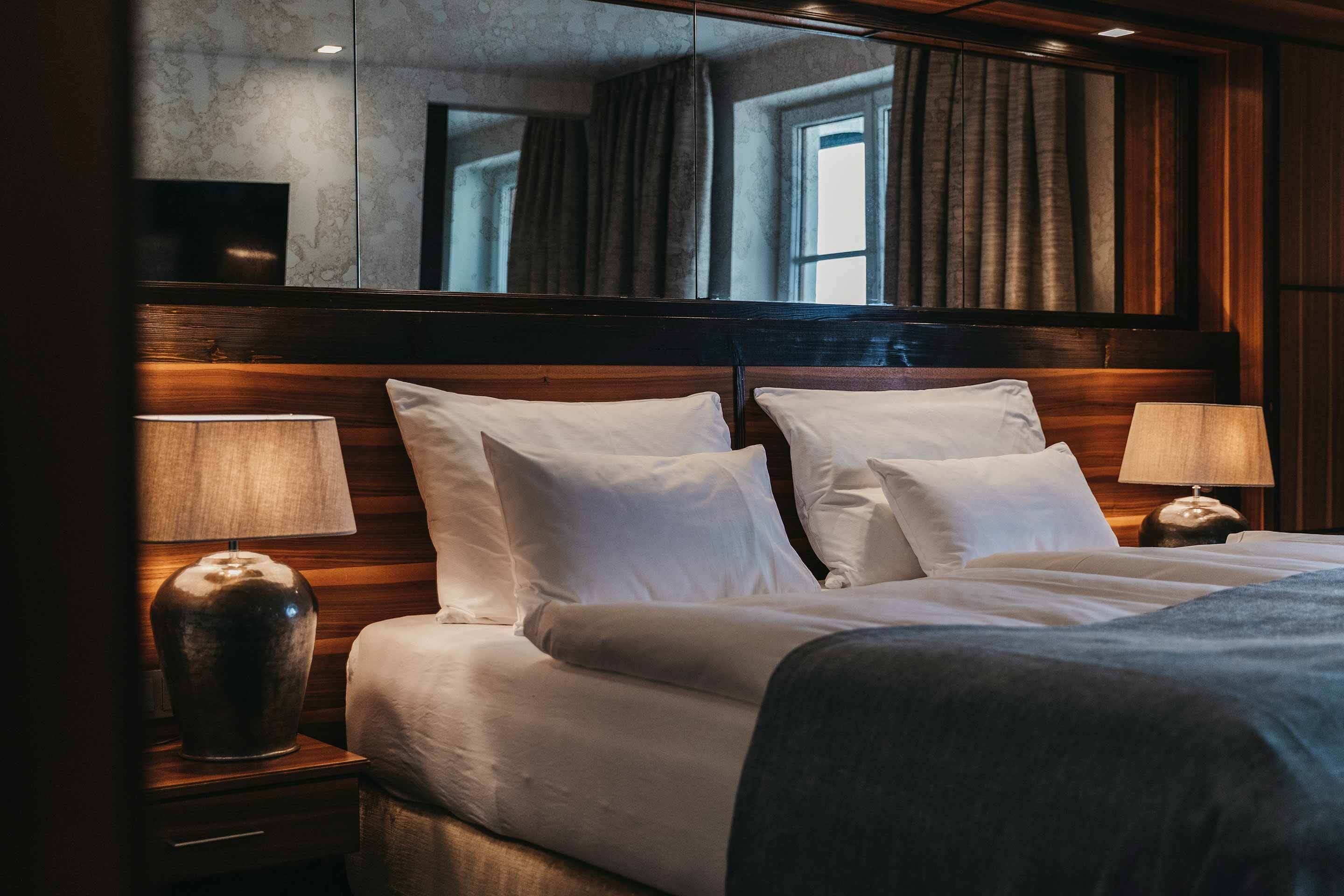 Una habitación de hotel elegante con una cama grande, dos mesitas de noche con lámparas, un espejo sobre la cama y vistas al exterior a través de una ventana.