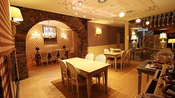 Comedor con pared de piedra, papel tapiz estampado y una araña. Mesas y sillas dispuestas para comer.