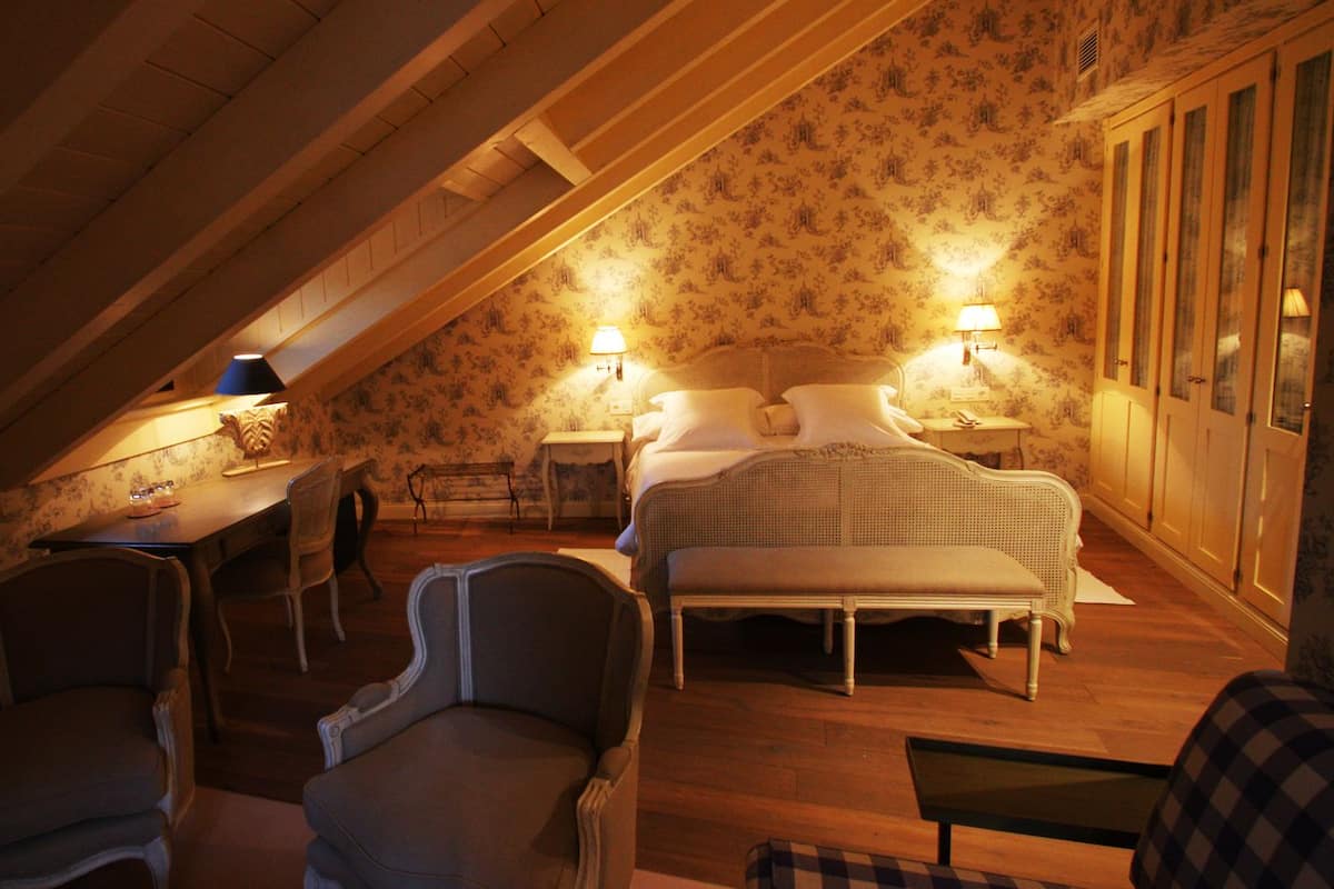 Ett mysigt sovrum på vinden med ett sluttande tak, en säng med vita sängkläder, två sänglampor och ett skrivbord i trä med en lampa.