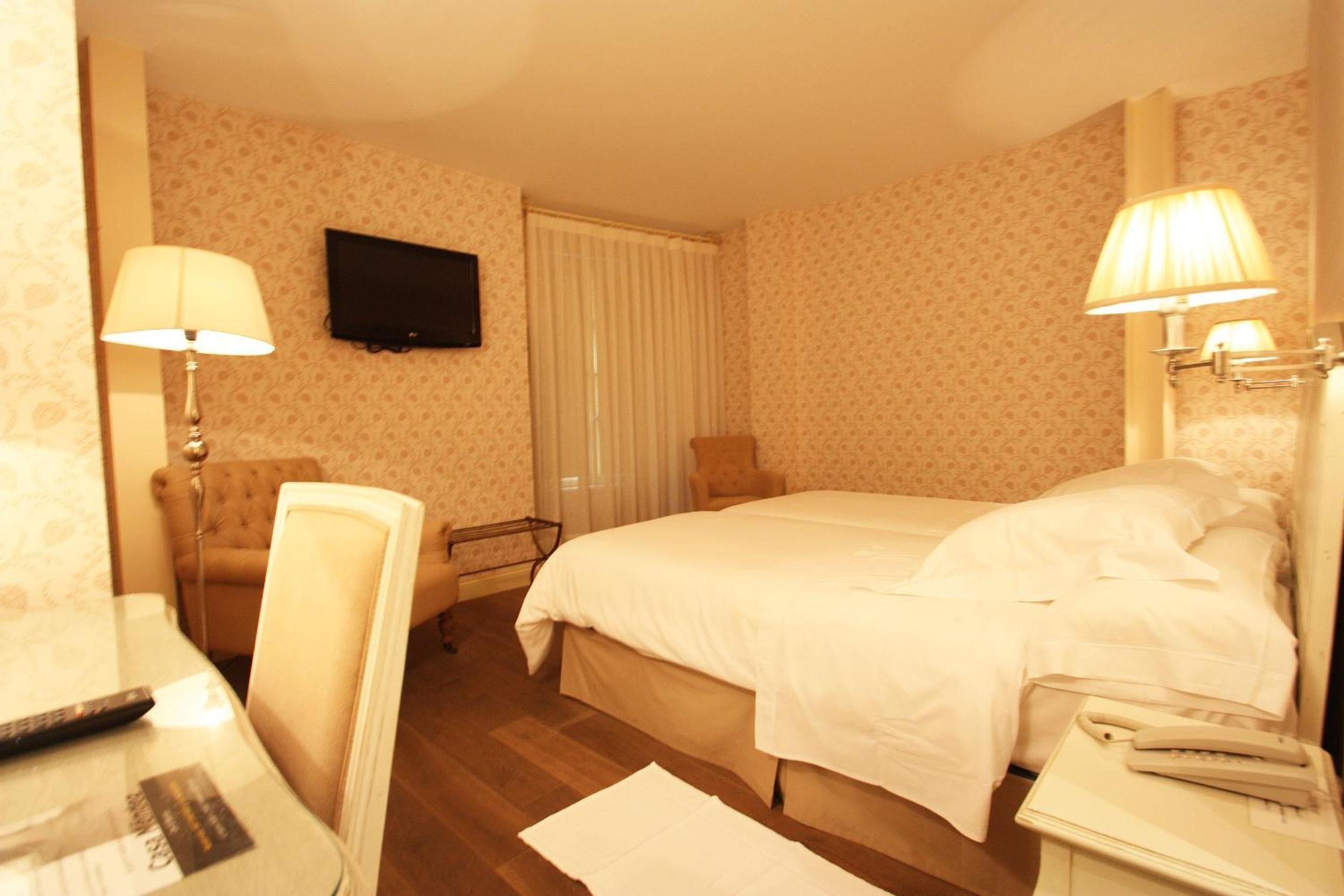 Habitación de hotel con cama, lámparas de noche, televisor montado en la pared, una silla y escritorio con control remoto.