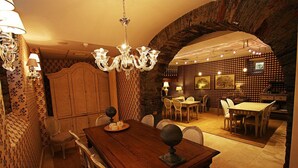 Un comedor con una mesa de madera, sillas y una araña. Se aprecia una pared con diseño y un arco de piedra en el fondo.