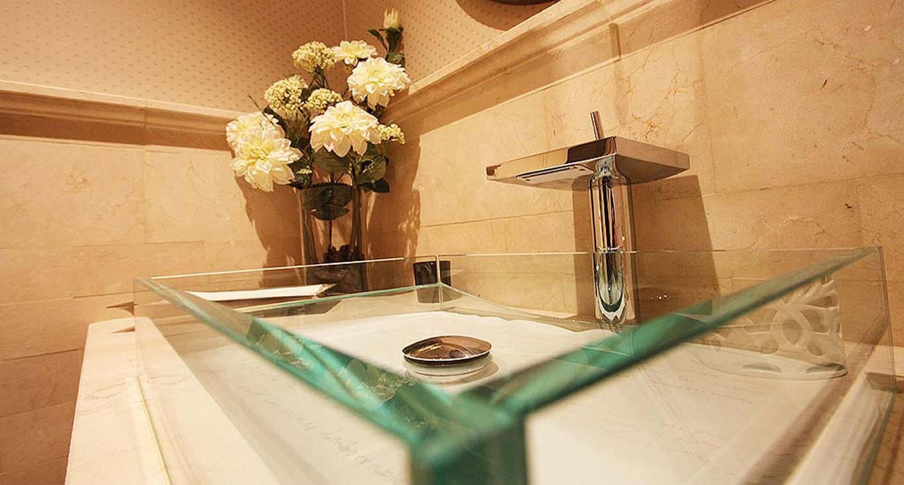 Un baño con lavamanos de vidrio, un espejo y un jarrón con flores en la pared.