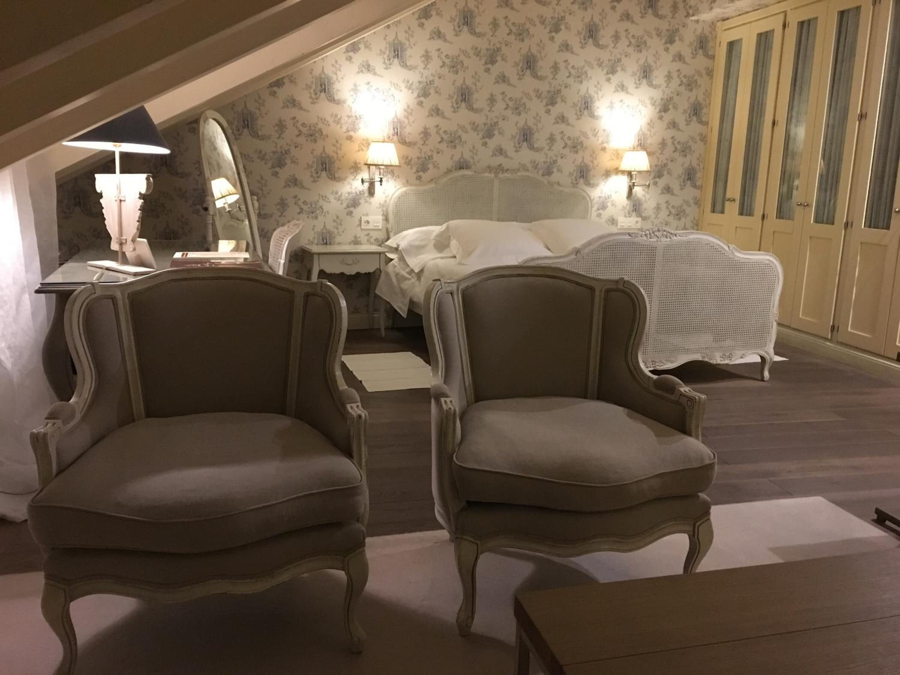 Un dormitorio con una cama, dos sillones, una mesita de noche, una lámpara y un espejo.