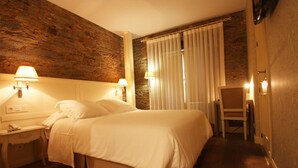 Una habitación de hotel con una cama grande, dos lámparas de noche, una silla y un ventanal con cortinas.