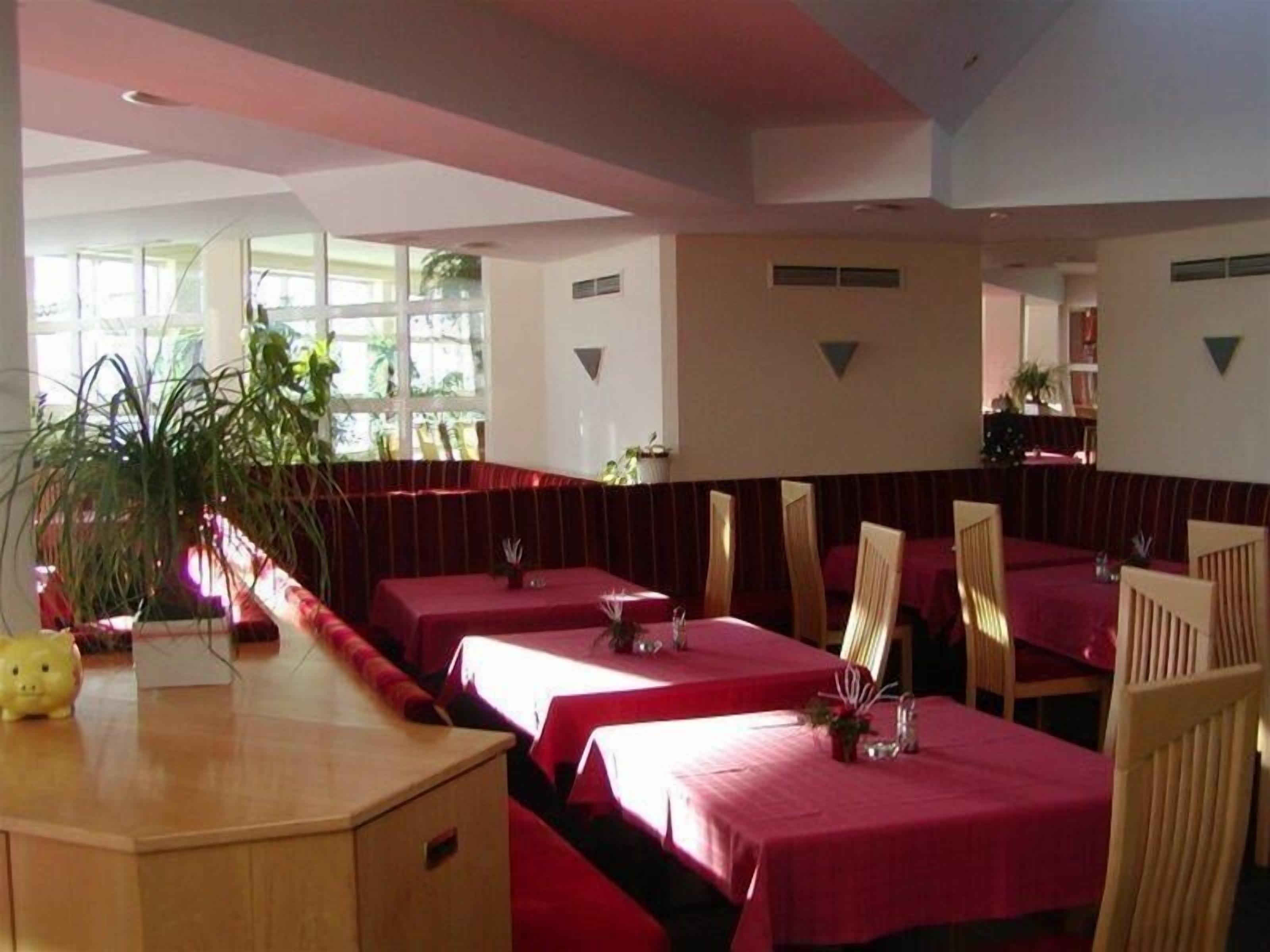 Een restaurant met rode tafelkleden en houten stoelen. Grote ramen zorgen voor veel natuurlijk licht.