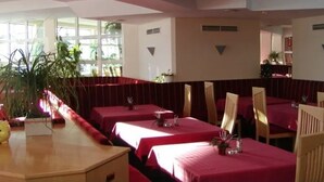 A restaurant with multiple tables covered in red tablecloths, wooden chairs, and potted plants. - Belmont (Imst)