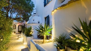 Un sendero estrecho bordeado por paredes blancas y plantas en macetas, que conduce a un edificio con una cúpula blanca y contraventanas negras.