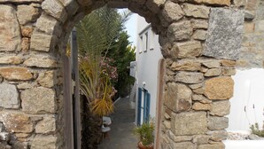 Un estrecho arco de piedra que conduce a un patio con plantas en macetas y un edificio blanco con contraventanas azules.