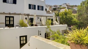 Un edificio blanco con contraventanas negras, un balcón con mesa y sillas, y vistas a una colina con otros edificios.