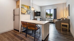 A modern hotel room with a kitchen area, a dining table with chairs, a sofa, and a TV. - Homewood Beaumont, TX (Beaumont)