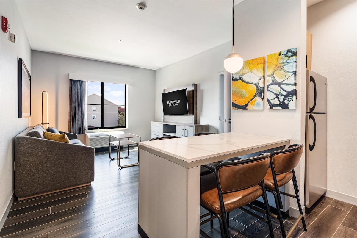 A modern kitchen with a central island, bar stools, a flat-screen TV, and a refrigerator.